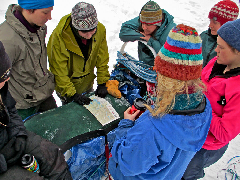 A group of people are gathered around what appears to be a sled or some equipment covered with a tarp and blue bags, possibly in a snowy environment. They seem to be examining a map or document. The individuals are wearing winter clothing, including hats and jackets, suggesting a cold climate. The scene implies they might be preparing for an outdoor activity or expedition.
